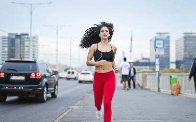 Woman jogging on a city street while listening to music with earphones, wearing red leggings and a black sports bra.