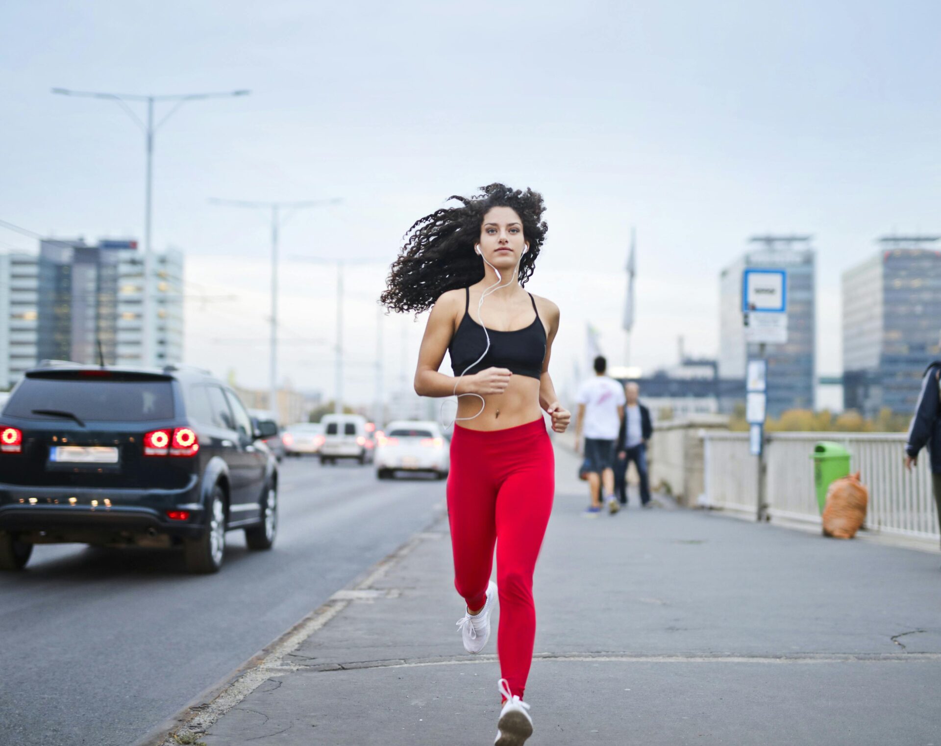 Woman jogging on a city street while listening to music with earphones, wearing red leggings and a black sports bra.
