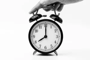 A hand holding a black alarm clock displaying the time at nearly 12 o'clock.