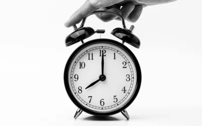 A hand holding a black alarm clock displaying the time at nearly 12 o'clock.